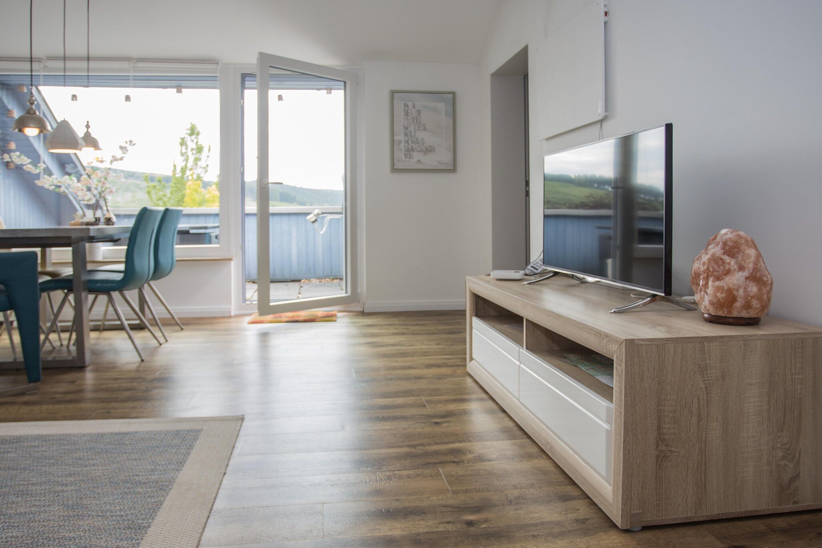 Una sala de estar moderna con suelo de madera, un televisor de pantalla plana sobre un mueble de madera, un ventanal con vistas al exterior y un comedor con sillas y una mesa.