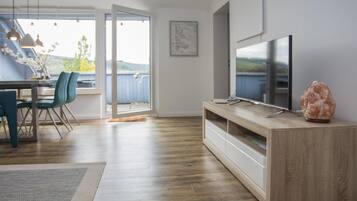 Un salon moderne avec un plancher en bois, un téléviseur à écran plat posé sur un meuble en bois, une grande fenêtre donnant sur l’extérieur et un coin repas avec des chaises et une table.