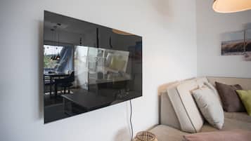 Ein modernes Wohnzimmer mit großem Flachbildfernseher an der Wand, einer Couch mit Kissen, einer Rattan-Stehlampe und einem Essbereich mit Tisch und Stühlen.
