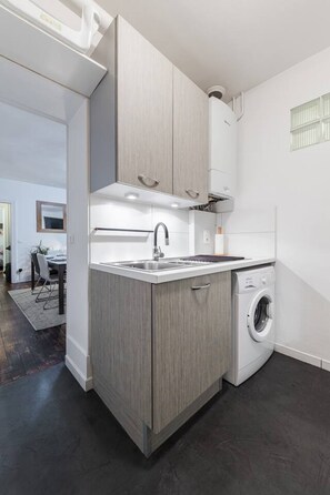 Une cuisine compacte dotée d’un lave-linge encastré, d’un évier et de meubles de rangement en bois clair.