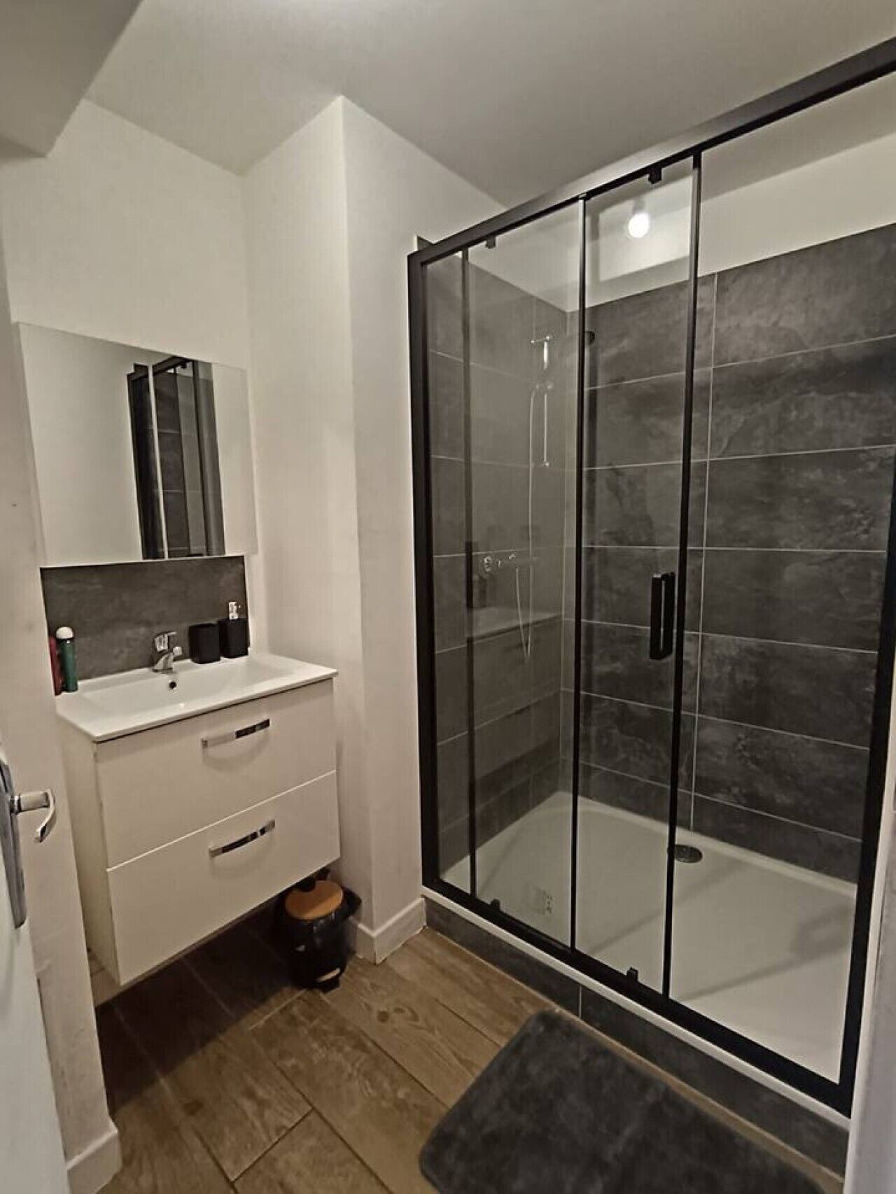 Un baño moderno con ducha de vidrio, tocador blanco con espejo y piso de madera.