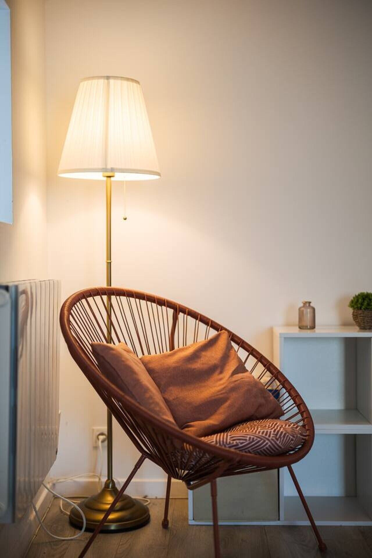 Ett modernt rum med en golvlampa, en flätstol med en kudde, ett element och en hylla med dekorativa föremål.