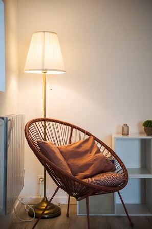 Ett modernt rum med en golvlampa, en flätstol med en kudde, ett element och en hylla med dekorativa föremål.