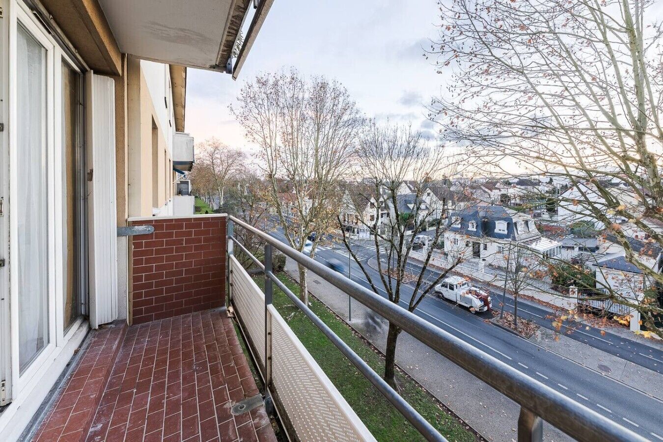 Un balcon avec un mur en briques, une rambarde en métal et une vue sur une rue résidentielle bordée de maisons et d’arbres.