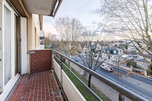 Un balcon avec un mur en briques, une rambarde en métal et une vue sur une rue résidentielle bordée de maisons et d’arbres.