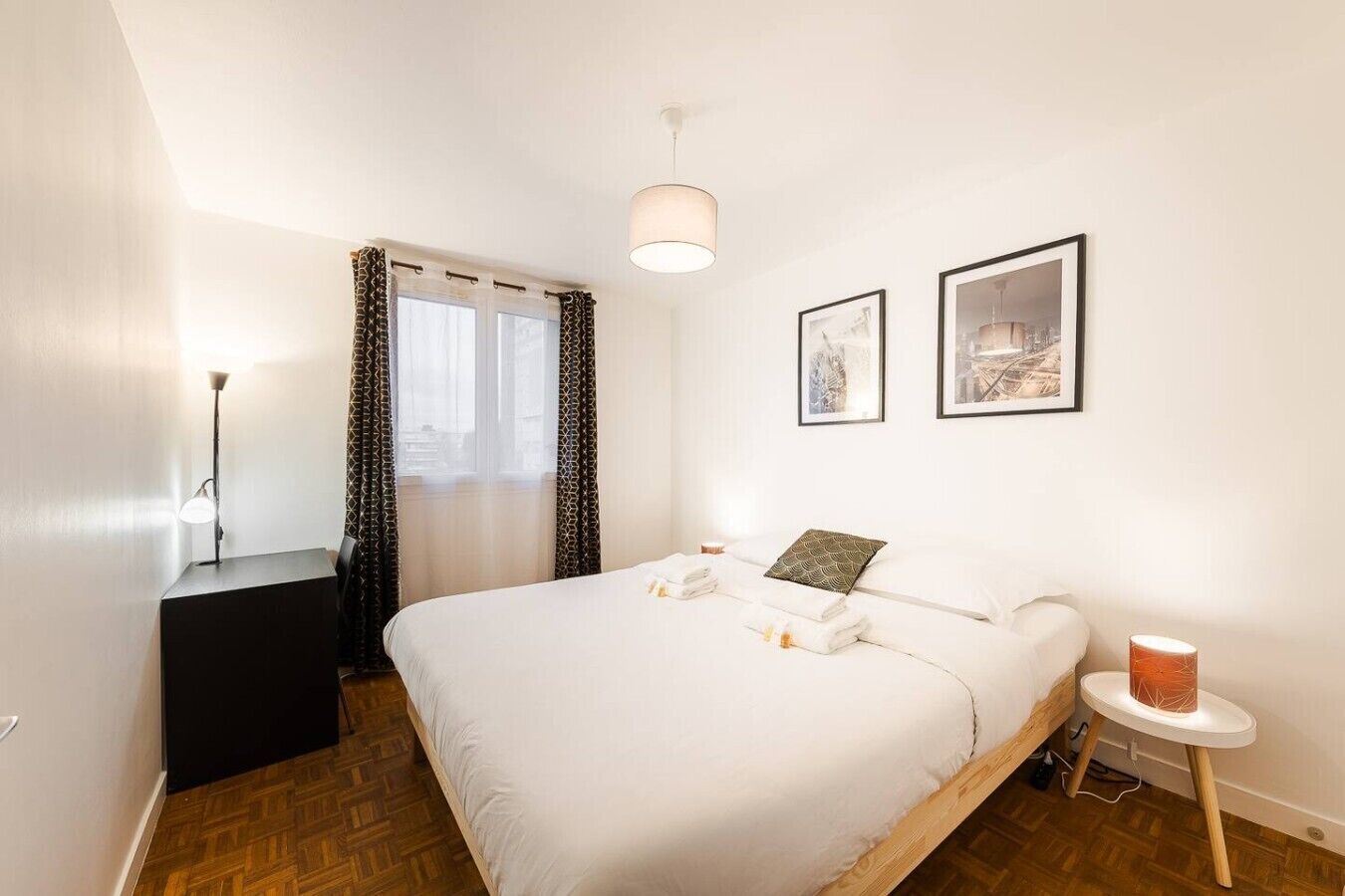 Ein Hotelzimmer mit Bett, Nachttisch, Lampe, einem Fenster mit Vorhängen und zwei gerahmten Bildern an der Wand.