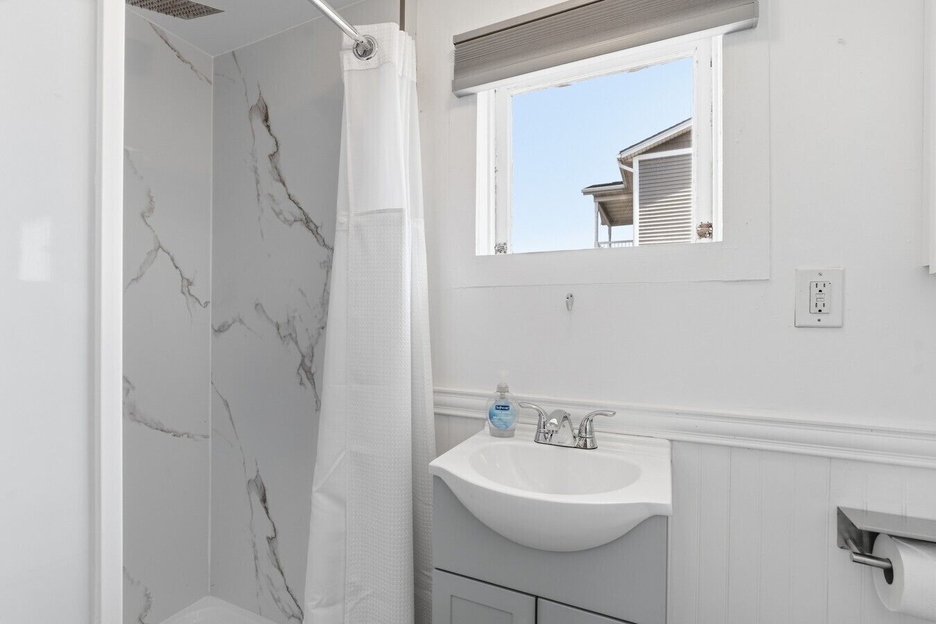 A bathroom with a marble shower wall, a white sink, and a window with a view of a building.