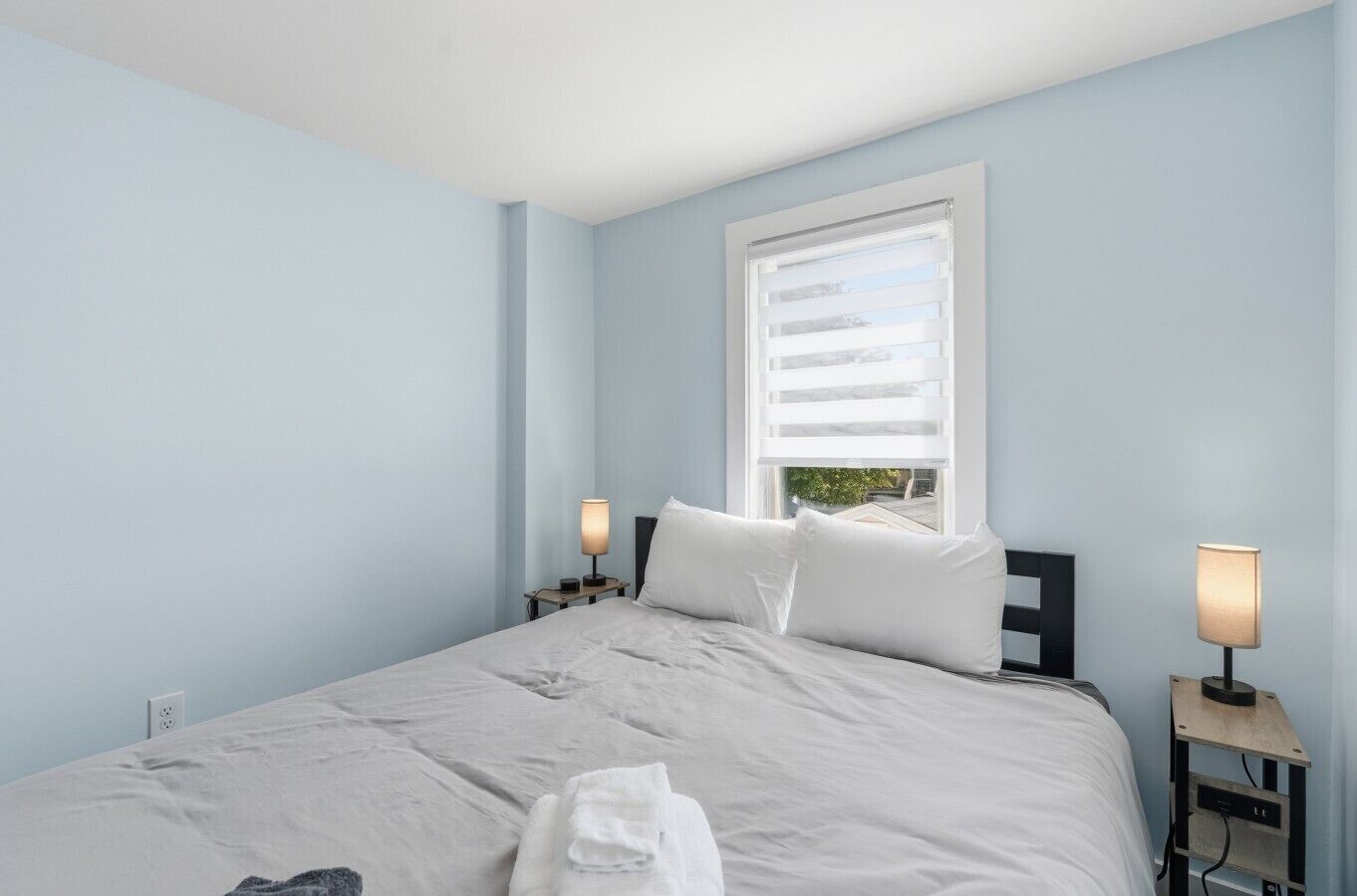 A bedroom with a bed, two bedside tables with lamps, a window with blinds, and a towel on the bed.