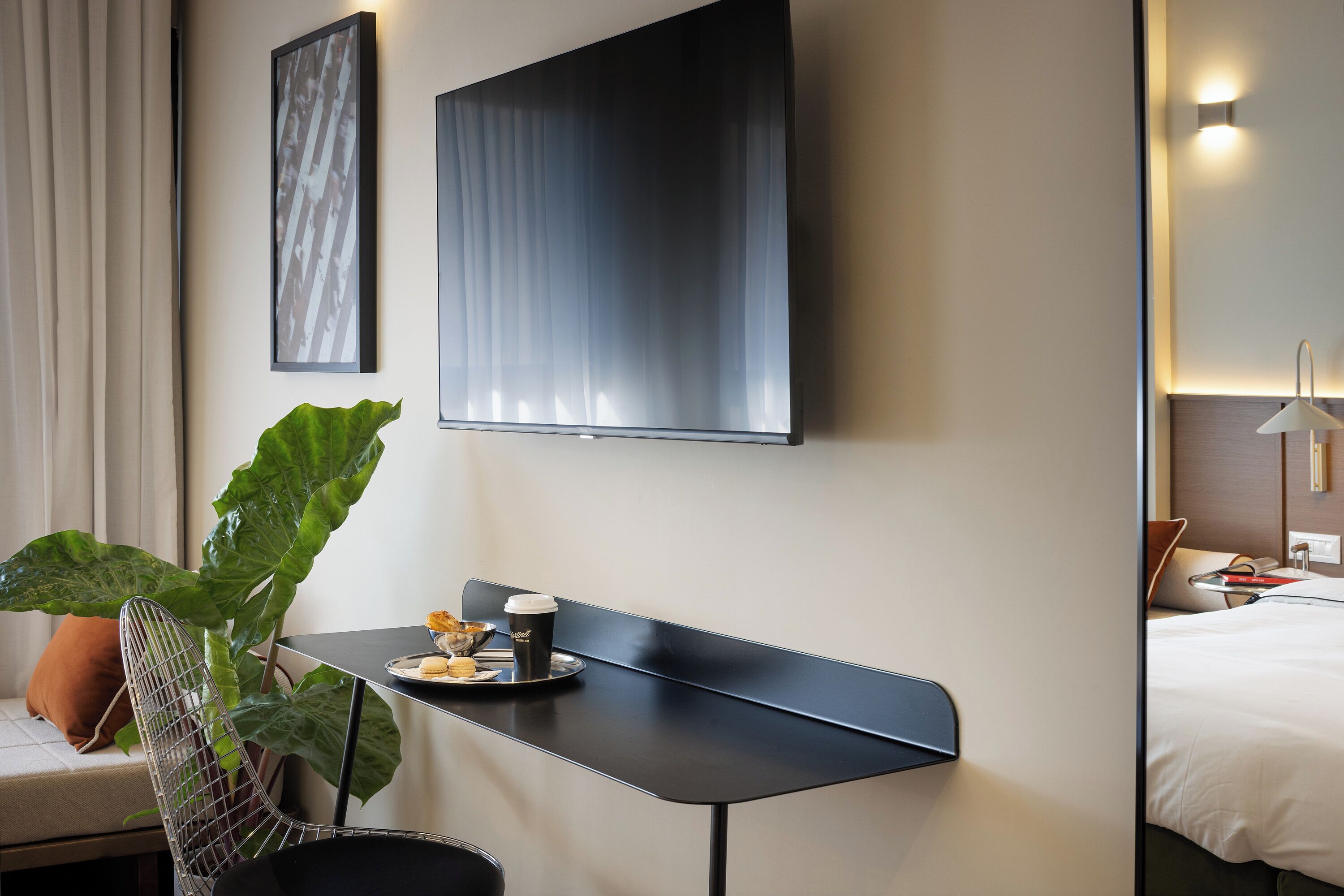 Habitación de hotel con cama, escritorio, taza y pasteles, una planta grande y televisor montado en la pared.