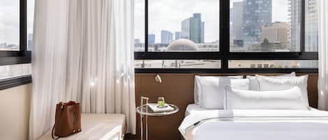 A hotel room with a bed, a small table with a drink, a handbag, and a view of a cityscape through large windows.