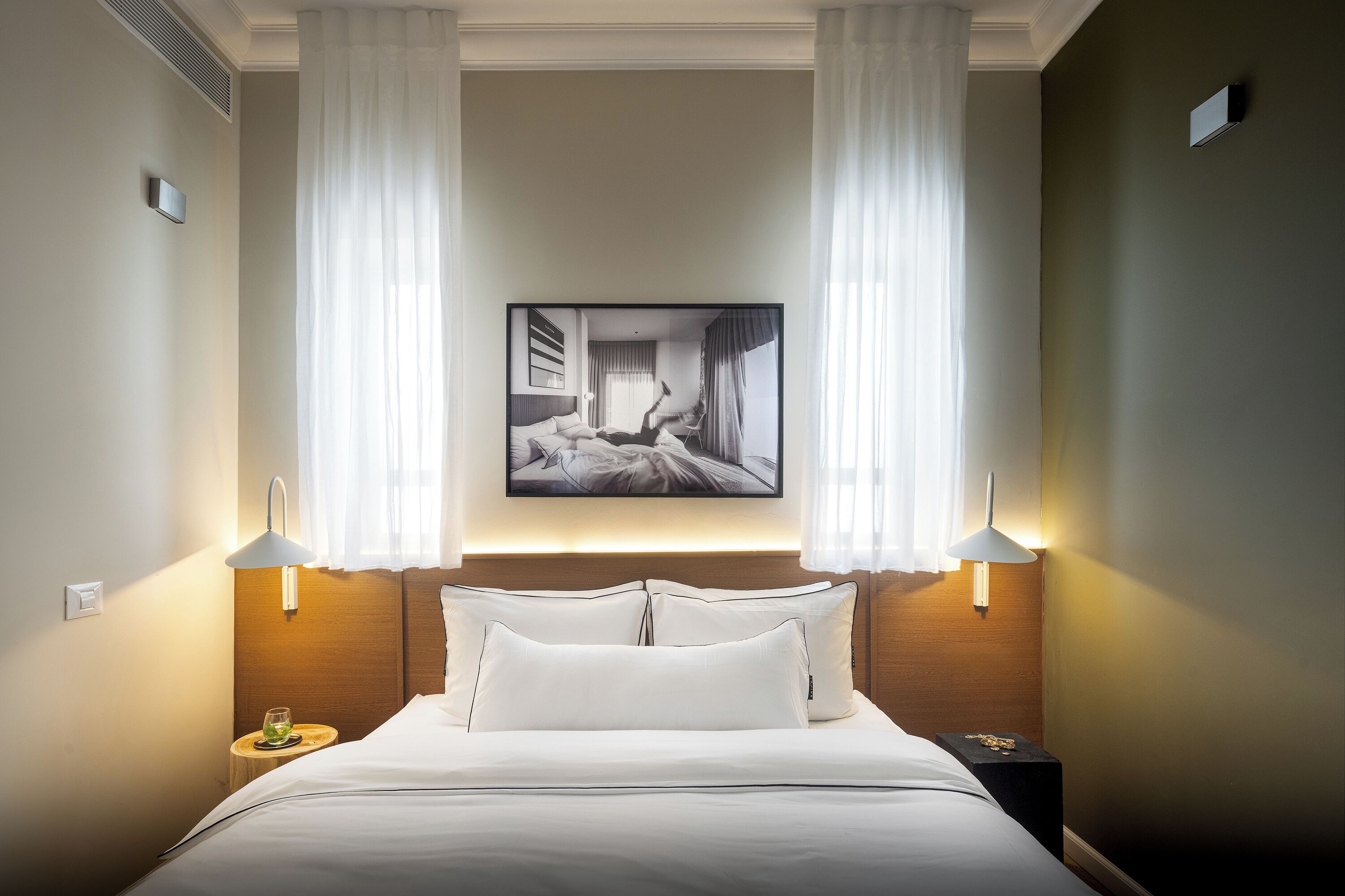A hotel room with a bed, two bedside lamps, a nightstand, and a framed photograph on the wall.