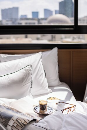 Una cama con una taza de café, un par de gafas y un plato de comida, con vista a la ciudad a través de una ventana.