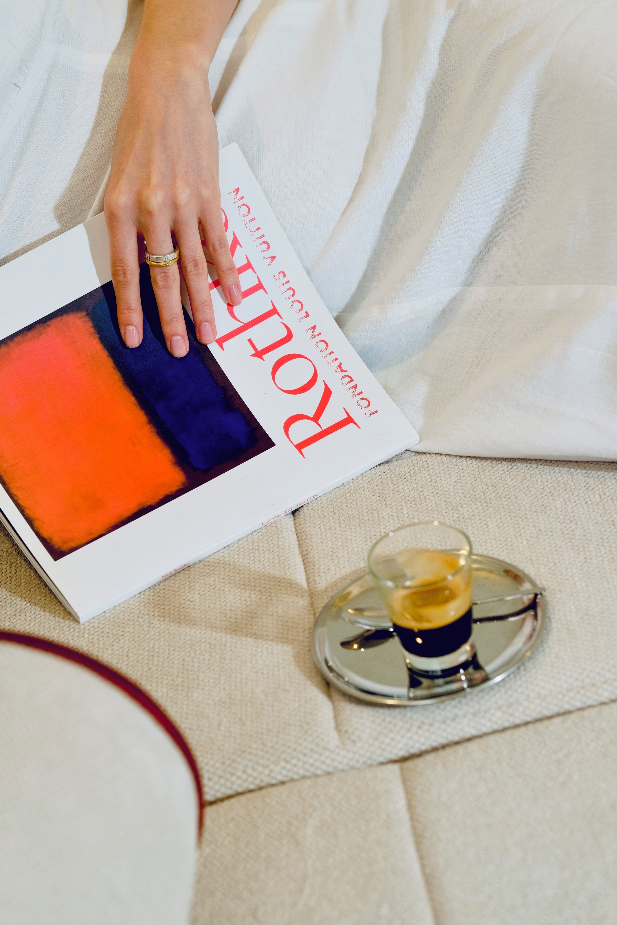 Una persona che tiene in mano una rivista con copertina rossa e blu, un bicchiere di un liquido scuro e un vassoio con una tazza.