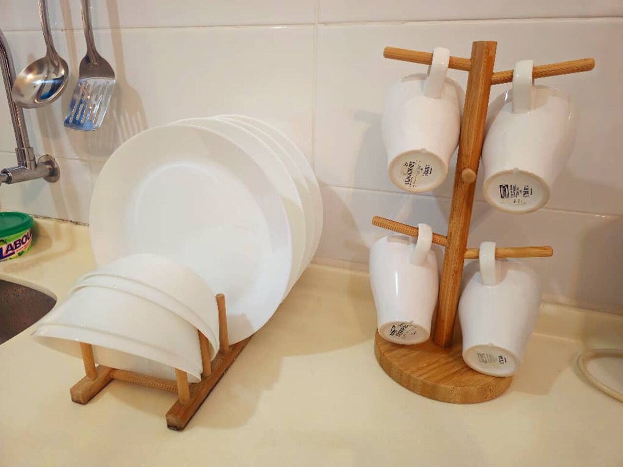 A kitchen counter with a dish rack holding plates and cups, a green container labeled 'LABOU', and a wooden spoon hanging on the wall.