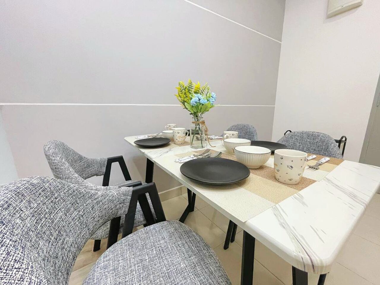 A dining area with a table set for four, chairs, a vase with flowers, and a wall-mounted light fixture.