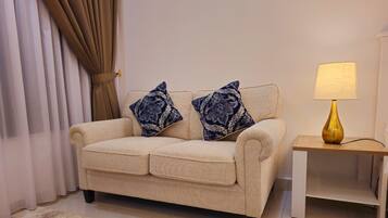 A beige sofa with two patterned pillows, a small white side table with a lamp, and a beige shag rug.