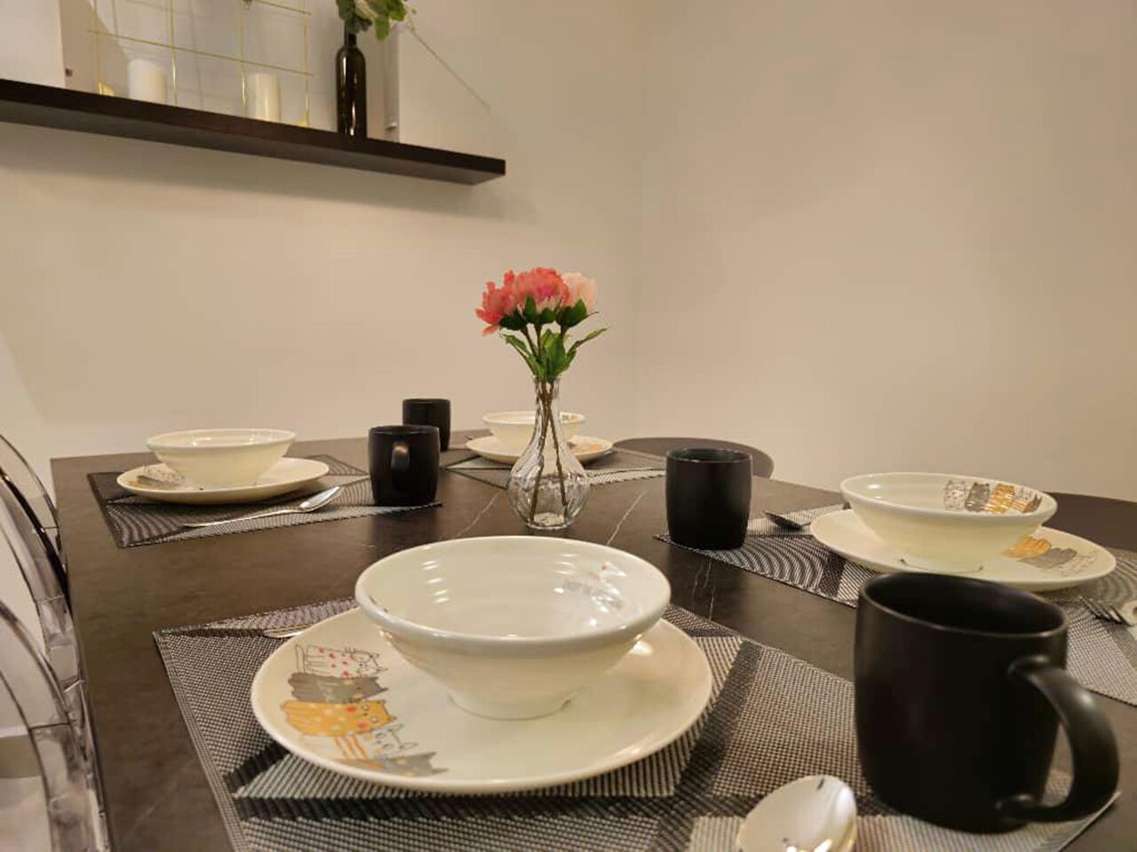 A dining table set with bowls, plates, a vase with flowers, and black mugs.