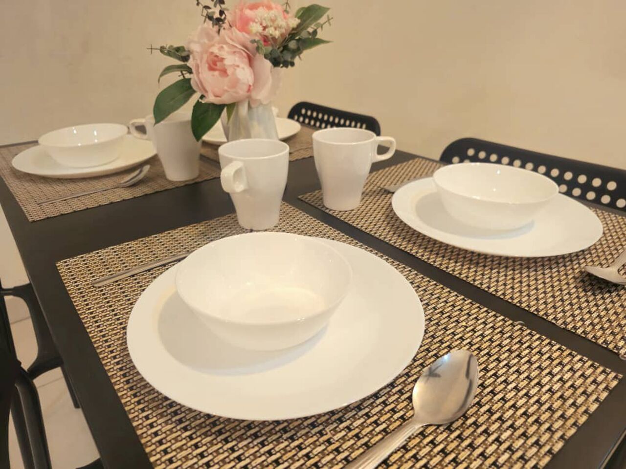 Una tavola imbandita con piatti, ciotole e tazze bianche, un sottopiattino intrecciato e un vaso di fiori.