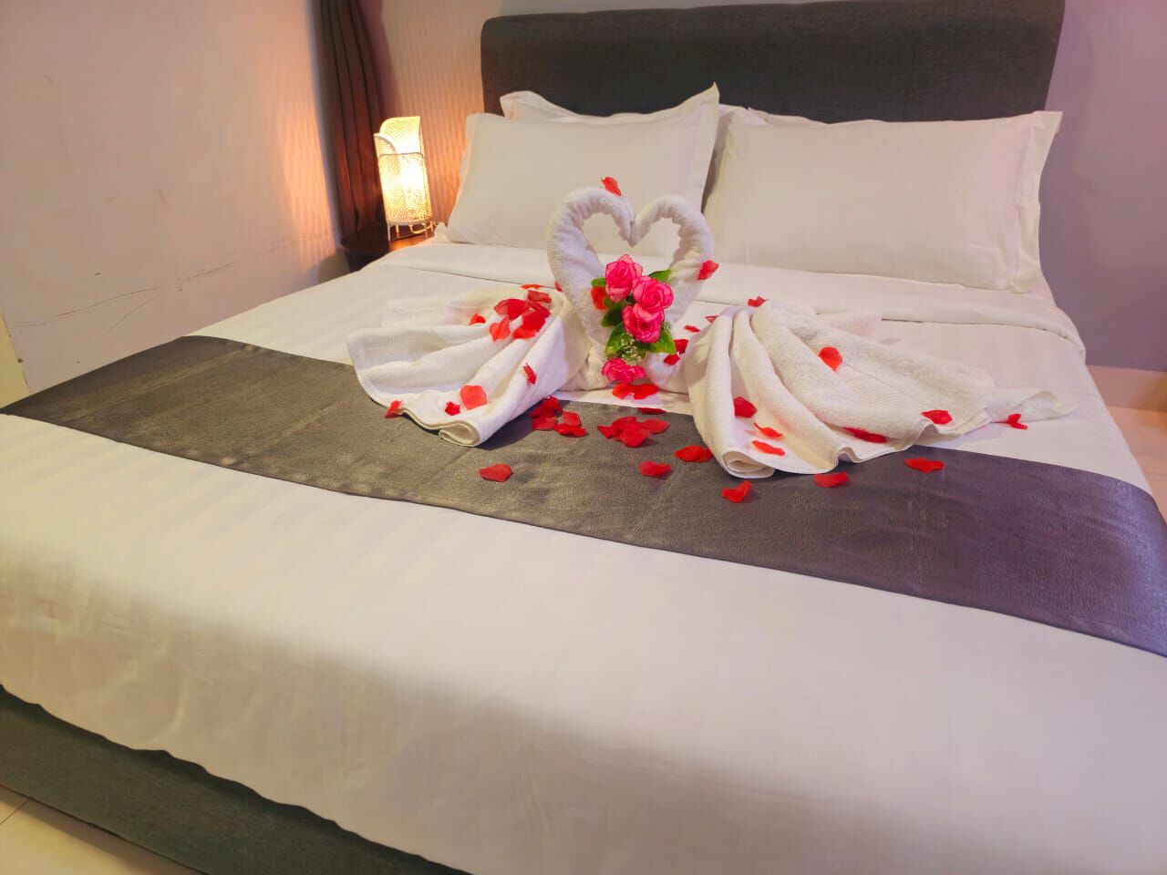 Una cama bien hecha con una toalla dispuesta en forma de corazón y pétalos de rosa, una lámpara de noche y un cabecero oscuro.