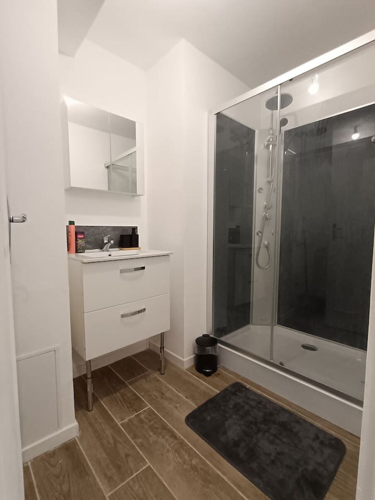 Un baño moderno con ducha, un tocador blanco con cajones, un espejo y un cesto de basura negro.