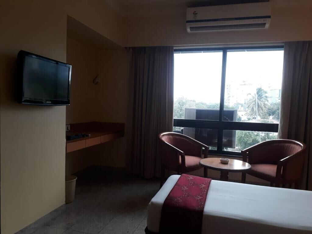 A hotel room with a bed, a television, a small table, two chairs, a window with curtains, and an air conditioning unit.