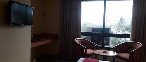 A hotel room with a bed, a television, a small table, two chairs, a window with curtains, and an air conditioning unit.