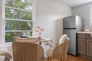 Eine Küche mit Kühlschrank, Korbstühlen, einem kleinen Tisch mit einer Vase und einem Fenster mit Blick auf Bäume.