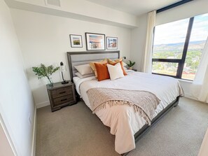 A bedroom with a bed, bedside tables, a nightstand, a window with a view, and framed pictures on the wall. - 2 Bedroom Lakeview in Downtown Kelowna with Rooftop Terrace + EV Parking (Kelowna)