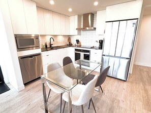 A modern kitchen with stainless steel appliances, a glass dining table, and white cabinetry. - 2 Bedroom Lakeview in Downtown Kelowna with Rooftop Terrace + EV Parking (Kelowna)