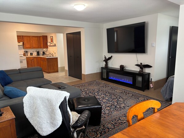 A living room with a sofa, ottoman, TV, entertainment unit, and a dining area with a table and chairs. - Campbell River discovery passage view guest suite (Campbell River)
