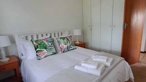 Un lit bien fait, avec du linge de lit blanc et des coussins aux motifs tropicaux, une table de chevet avec une lampe et une armoire en bois dans une pièce aux murs de couleur claire.