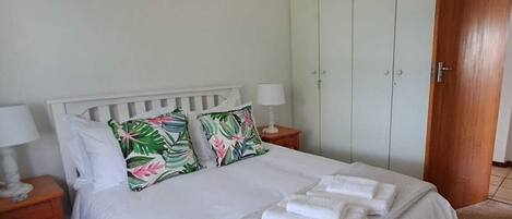 A neatly made bed with white linens and tropical pillows, a bedside table with a lamp, and a wooden wardrobe in a room with light-colored walls.