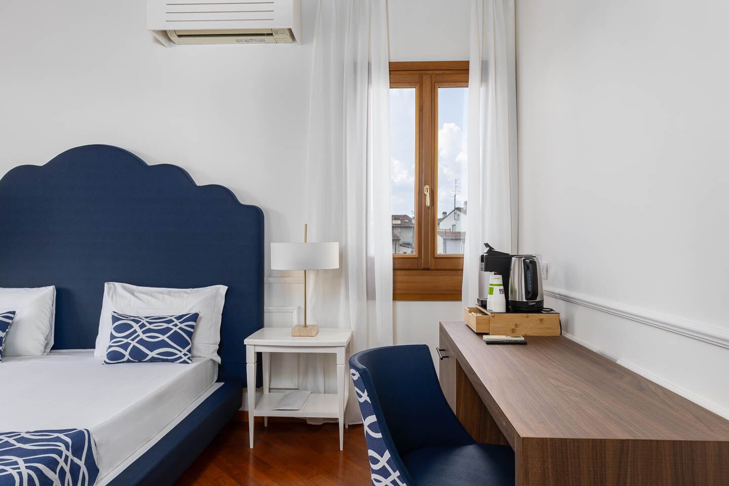 A hotel room with a bed, bedside table, lamp, desk, chair, and a window with a view.