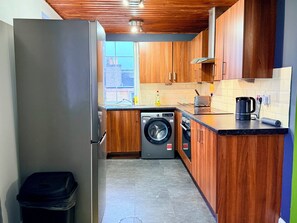 A compact kitchen with wooden cabinets, a washing machine, a coffee maker, and a trash bin. - Central 4Bed, Caledonian Stay, Haymarket, Sleeps 8 (Edinburgh)