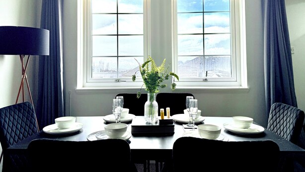 Ein Esszimmer mit einem für vier Personen gedeckten Tisch, zwei mit Vorhängen versehenen Fenstern und einer Stehlampe.