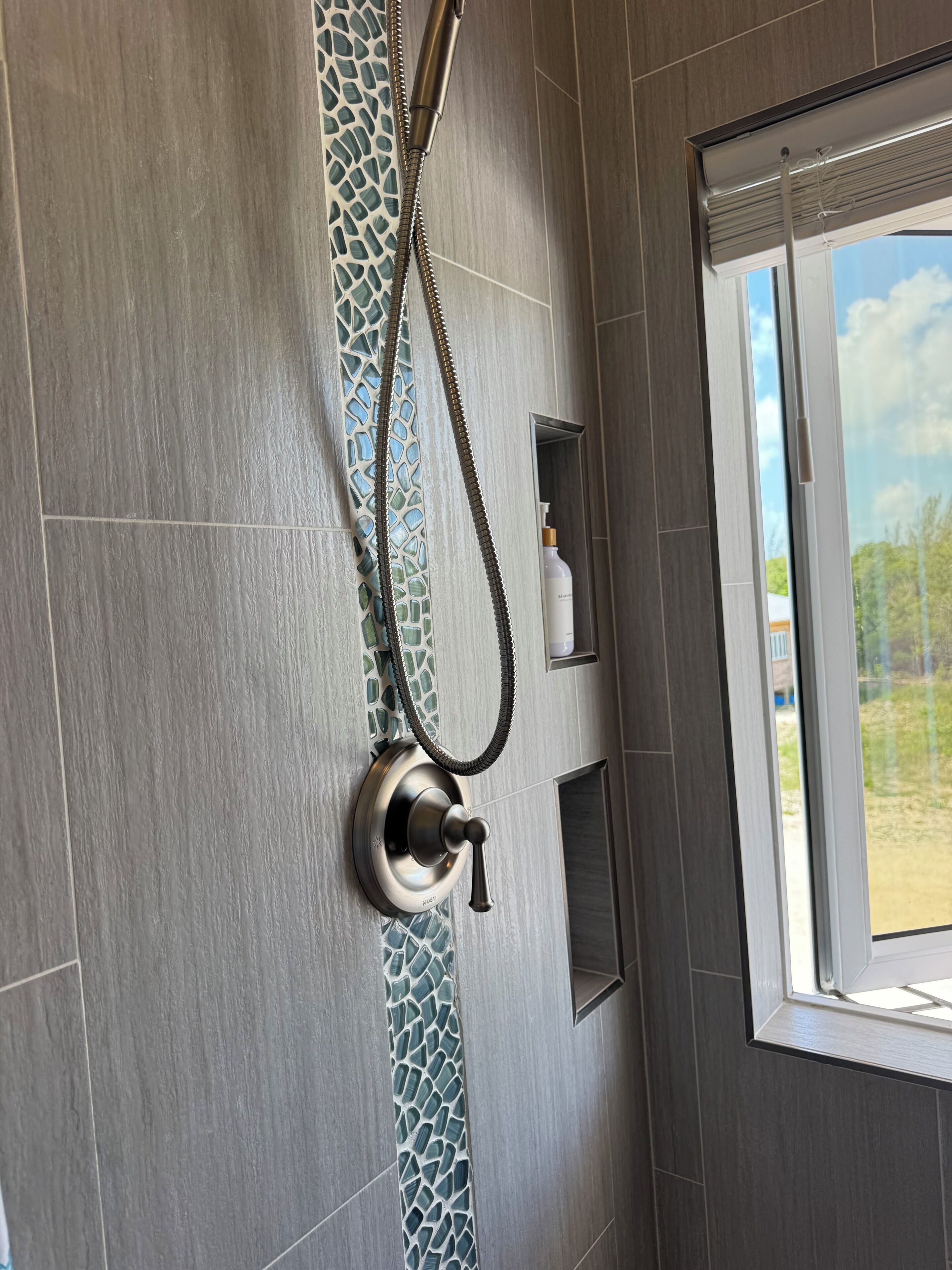 Ducha con ducha de mano, dispensador de jabón y ventana con vista a follaje.
