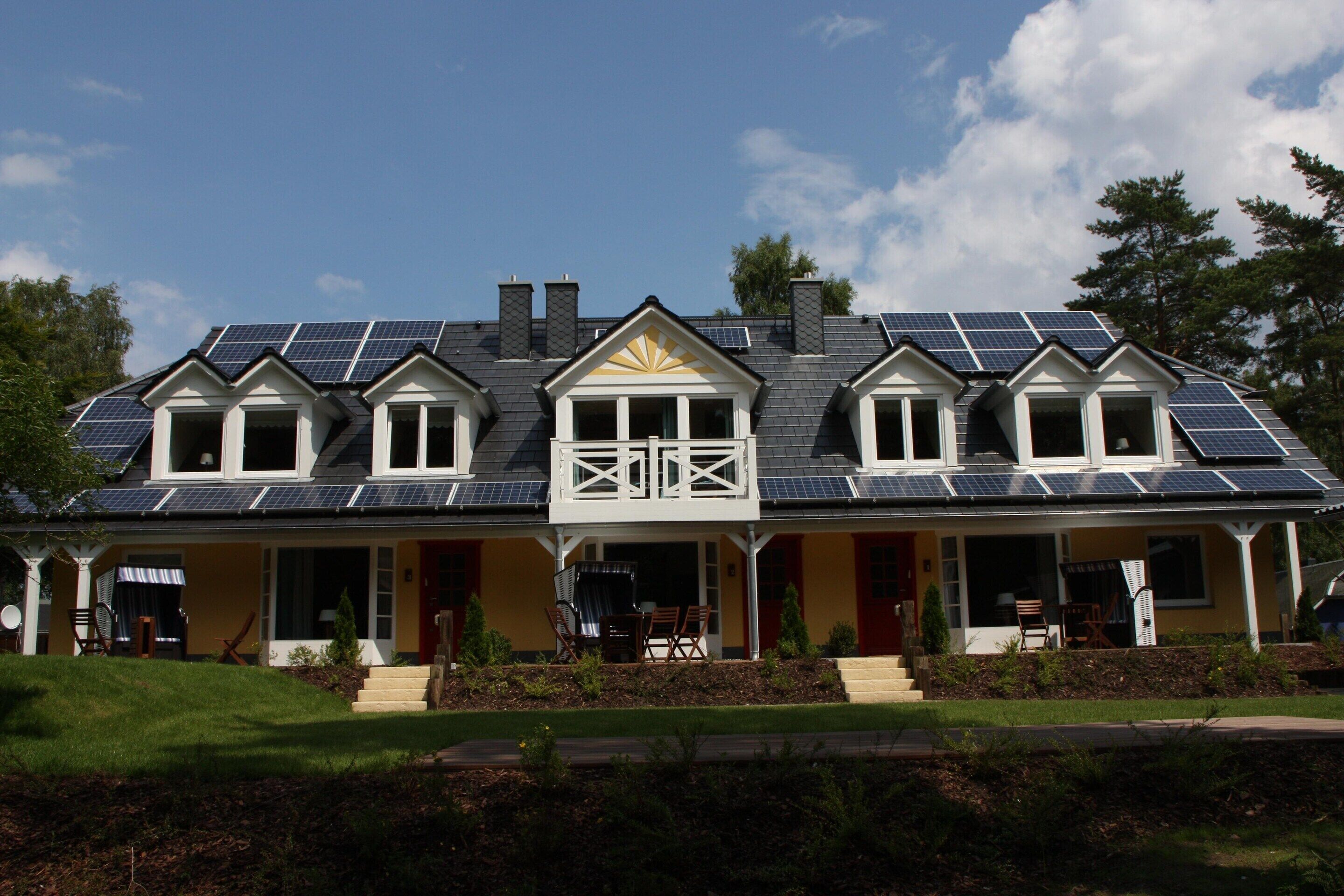 A two-story house with solar panels on the roof, a covered porch, and a front yard with a lawn and shrubs.