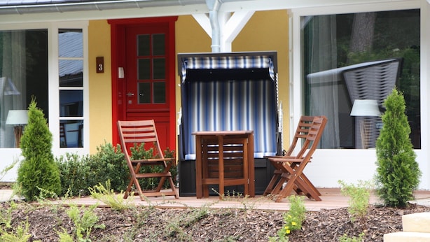 Et hus med en rød dør, et lille bord, to træstole og et vindue med gardin.