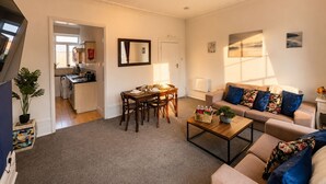A living room with a sofa, coffee table, and dining table. There is a kitchen in the background. - Spacious 3BR Camden Home, Sleeps 6, Fast Wi-Fi, Walk to Tube & Markets! (London)