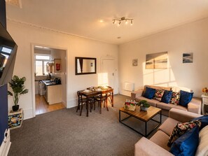 Ein Wohnzimmer mit einer Couch, einem Couchtisch und einem Esstisch. Im Hintergrund ist eine Küche zu sehen.