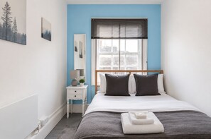 Ein Schlafzimmer mit Bett, Nachttisch, Lampe und einem Fenster mit Jalousien.