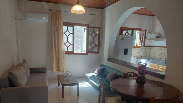 A living room with a sofa, a table, and a dining area with chairs. There is a window with curtains, a ceiling fan, and a view of the kitchen through an archway. - Antistoni Cozy  house in beautiful Agros with AC (Agros)