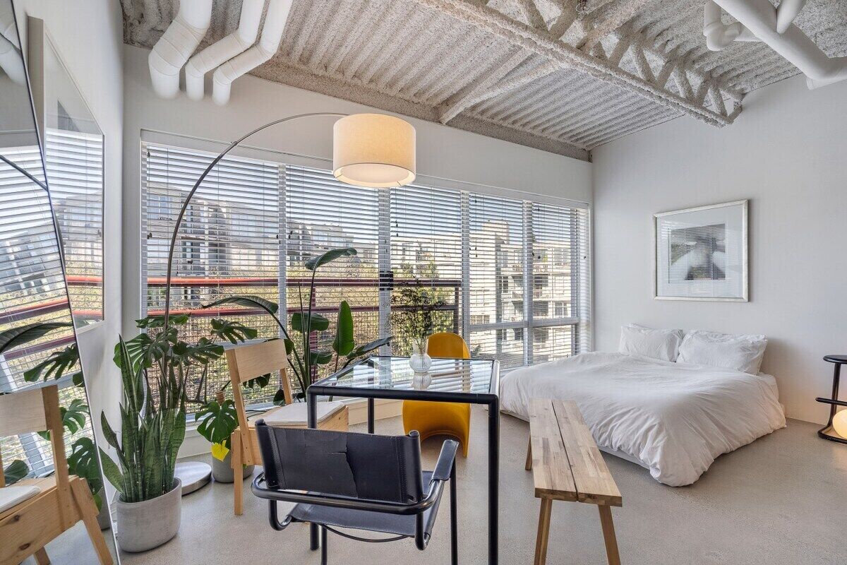 A modern bedroom with a bed, a glass table, and chairs, large windows with blinds, and a view of the city.