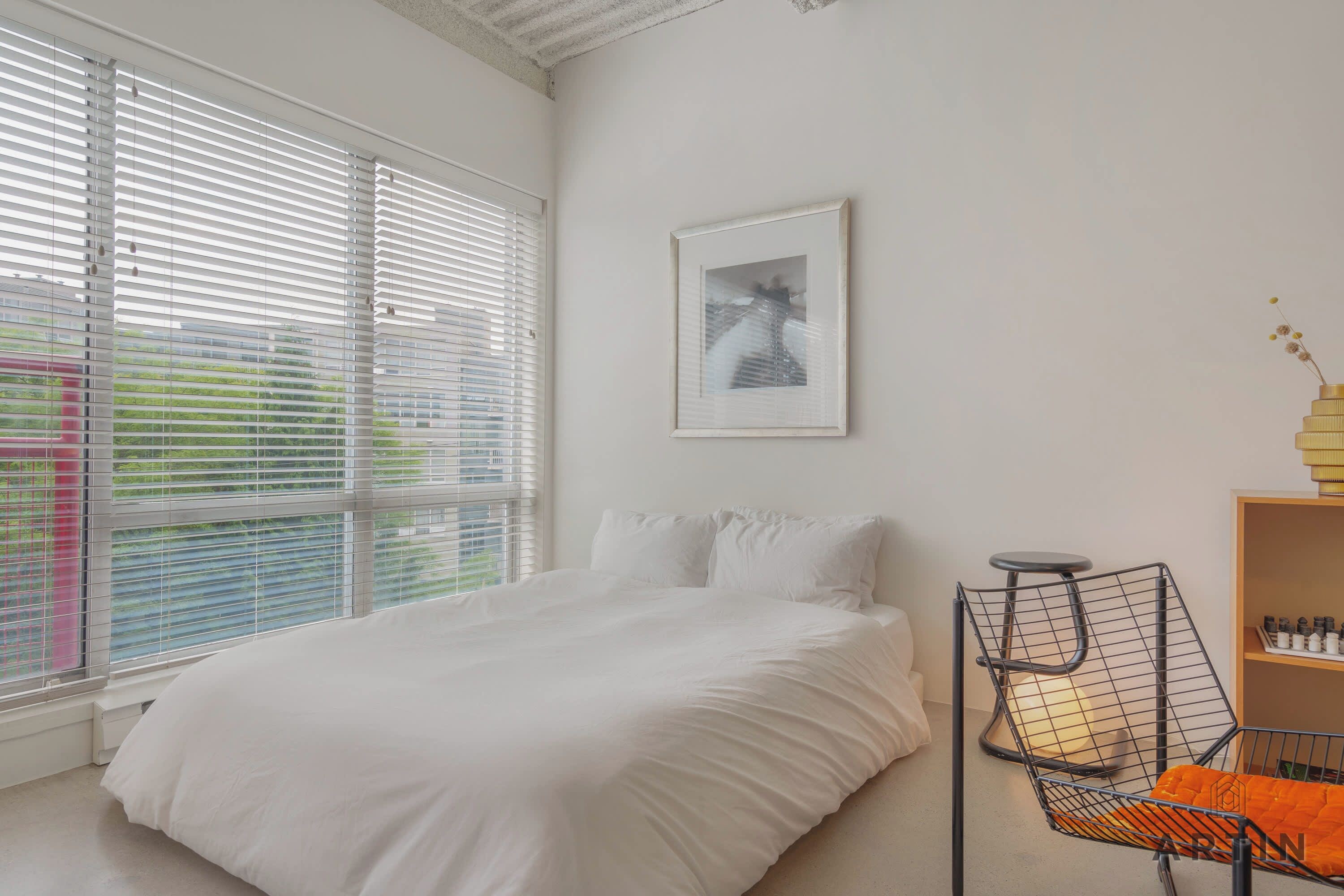 A bedroom with a bed, a chair, a framed picture, and a window with blinds.