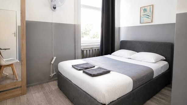 Un dormitorio con cama, una silla, un ventilador de pared, una ventana y un radiador.