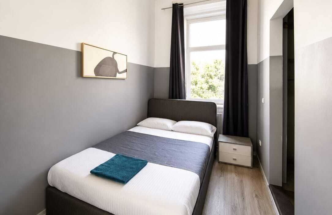 Un dormitorio con cama, mesita de noche, ventana con cortinas e imagen colgada en la pared.