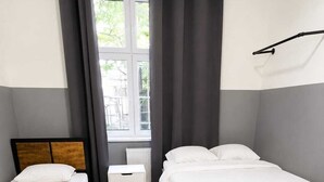 Una habitación con dos camas individuales, una silla blanca pequeña y un ventanal con cortinas.