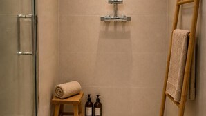 Ein modernes Badezimmer mit Duschkabine aus Glas, einem Holzschemel, zwei Shampooflaschen, einer Holzleiter und einem gewebten Korb mit Handtüchern.