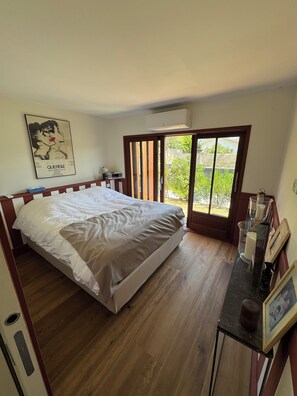 Une chambre à coucher comprenant un lit, un bureau avec un cadre photo, un climatiseur fixé au mur et une porte-fenêtre coulissante donnant sur un balcon.
