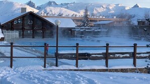 En træhytte med snedækket tag, en stenvæg og udsigt til fjelde i baggrunden.